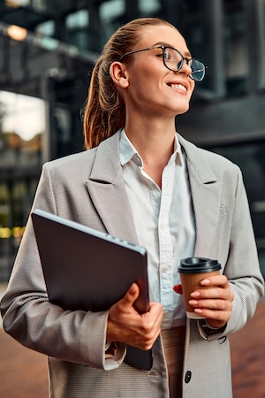Beautiful confident successful modern business woman wearing glasses holding a laptop and coffee and looking to the side while standing outside in the evening behind a dark blurred background.